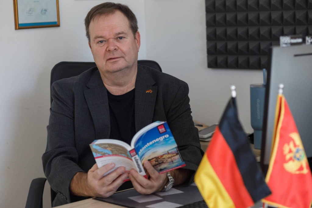 Michael Bader, Montenegro-Experte, am Schreibtisch mit einem aufgeschlagenen Montenegro-Reiseführer und der deutschen sowie montenegrinischen Flagge