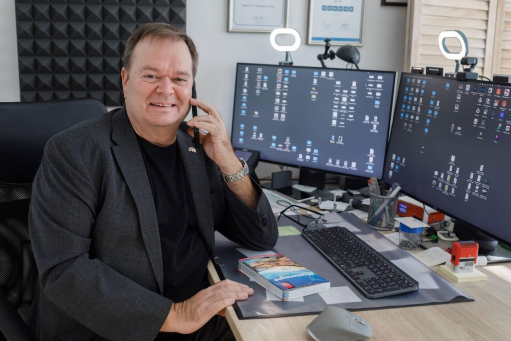 Michael Bader, Montenegro-Experte, telefoniert lächelnd an seinem Büroschreibtisch mit Montenegro-Reiseführer und zwei Monitoren